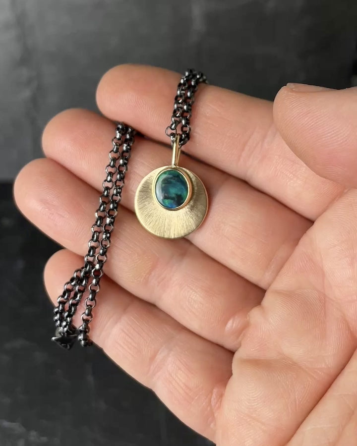 A hand is holding an Australian Black opal and Gold pendant that hangs on an oxidised sterling silver chain. The movement in the video shows off the the blue, green, and black swirls in the opals as well as the intricate engraved detail of the pendant.