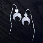 Lunar Thread Earrings on a black wooden background. These sterling silver earrings feature a textured disc and crescent moon hanging from a hook and fine chain fitting.