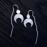 Lunar Thread Earrings on a black wooden background. These sterling silver earrings feature a textured disc and crescent moon hanging from a hook and fine chain fitting.