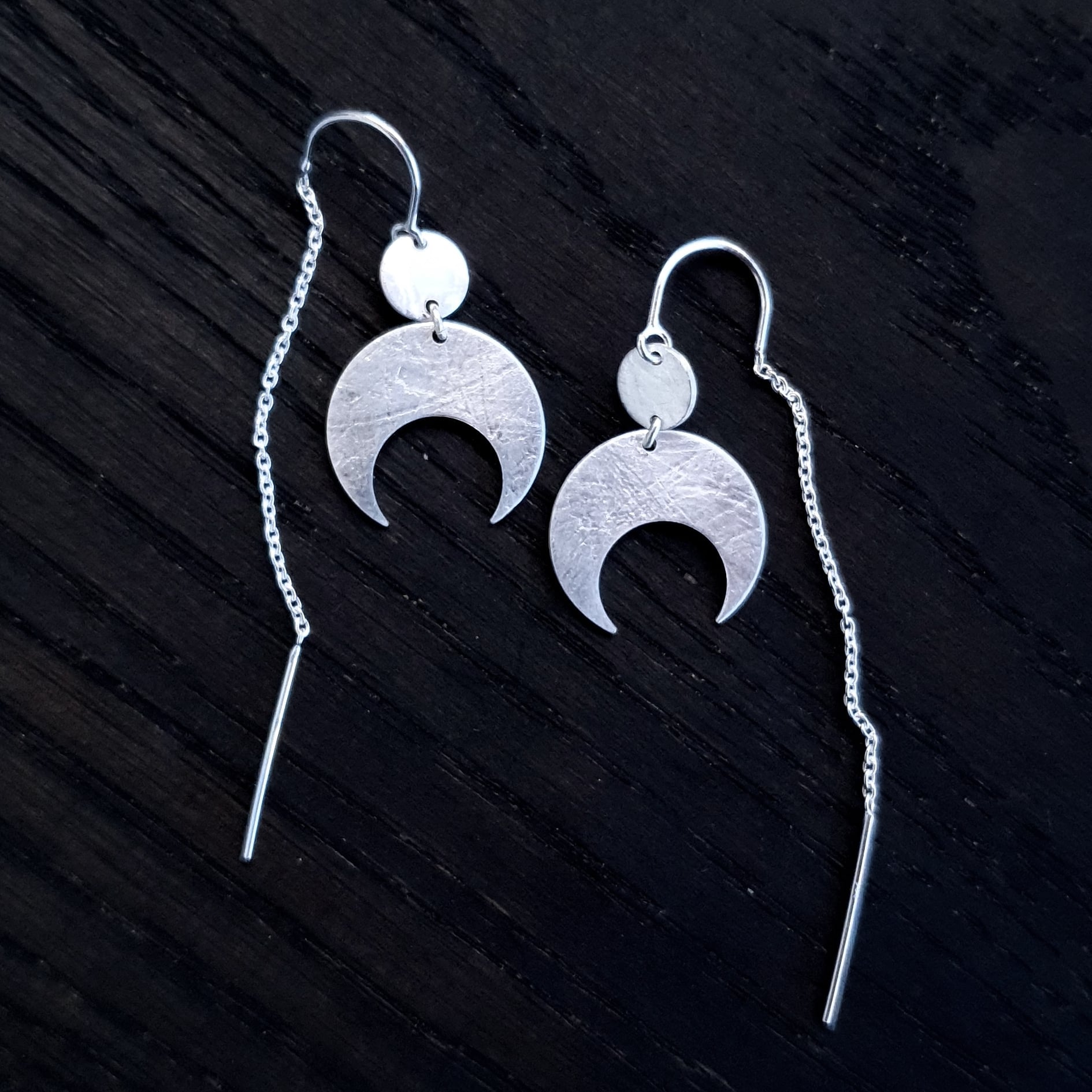 Lunar Thread Earrings on a black wooden background. These sterling silver earrings feature a textured disc and crescent moon hanging from a hook and fine chain fitting.