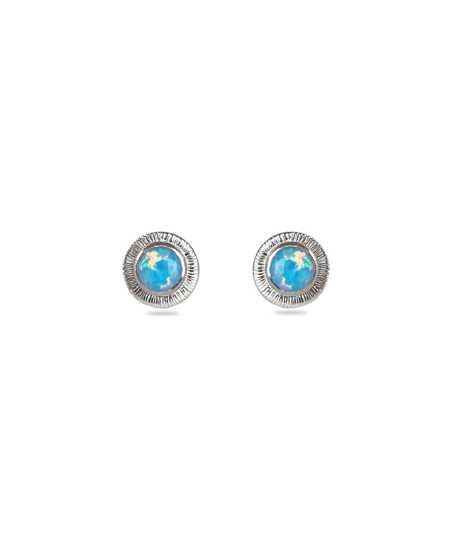 A pair of handmade opal stud earrings by Emily Eliza Arlotte, featuring light blue opals and an engraved textured halo, on a white background.