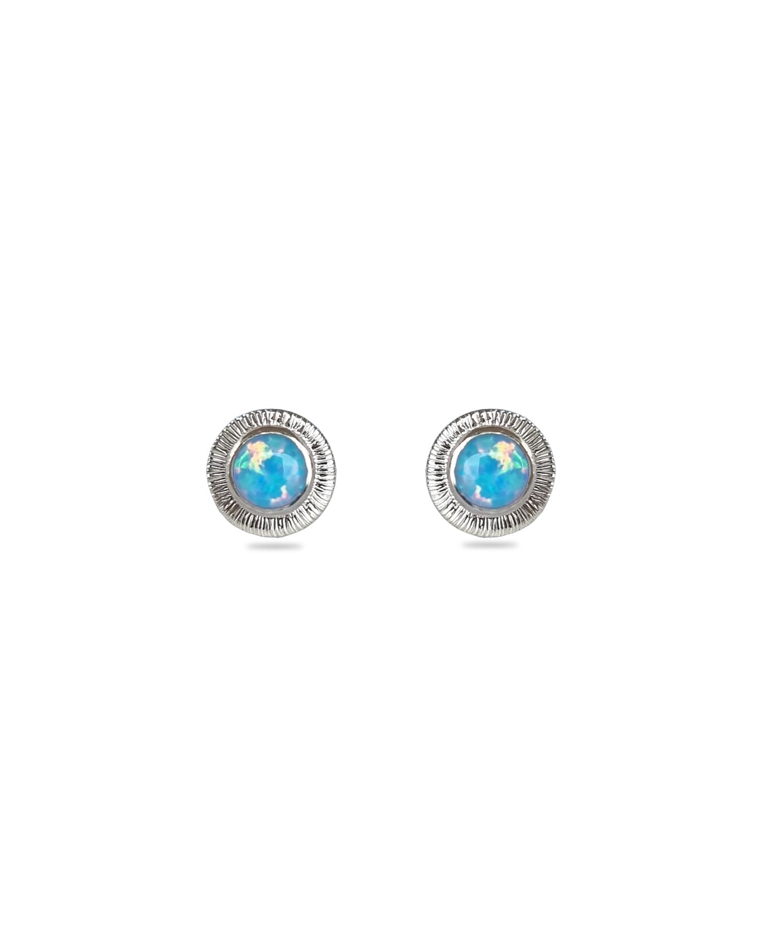 A pair of handmade opal stud earrings by Emily Eliza Arlotte, featuring light blue opals and an engraved textured halo, on a white background.