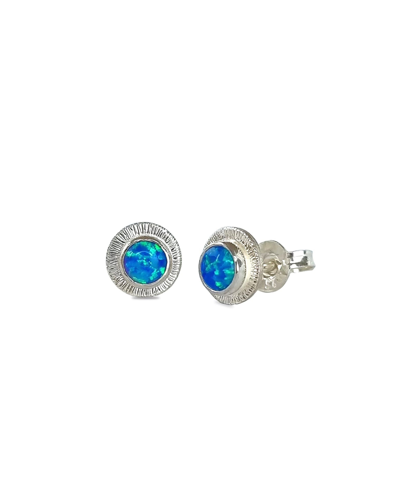 A pair of handmade opal stud earrings by Emily Eliza Arlotte, featuring a blue opals and an engraved textured halo, on a white background.