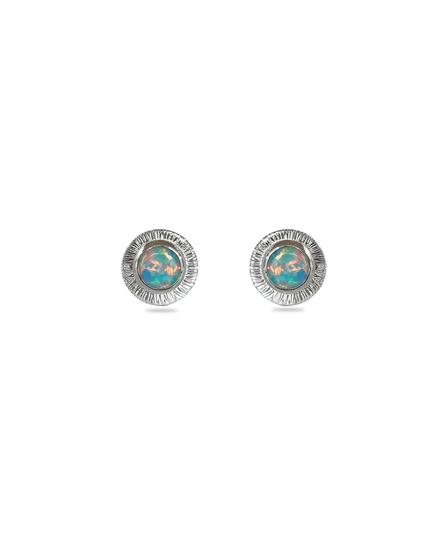 A pair of handmade opal stud earrings by Emily Eliza Arlotte, featuring mint green blue opals and an engraved textured halo, on a white background.