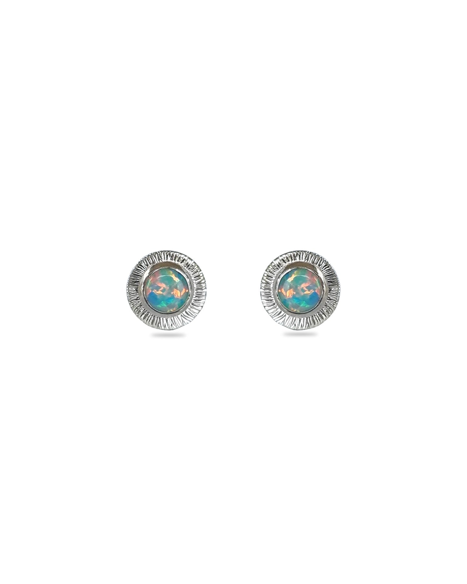 A pair of handmade opal stud earrings by Emily Eliza Arlotte, featuring mint green blue opals and an engraved textured halo, on a white background.