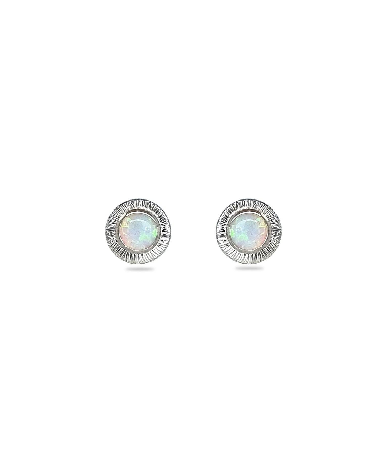A pair of handmade opal stud earrings by Emily Eliza Arlotte, featuring white opals and an engraved textured halo, on a white background.