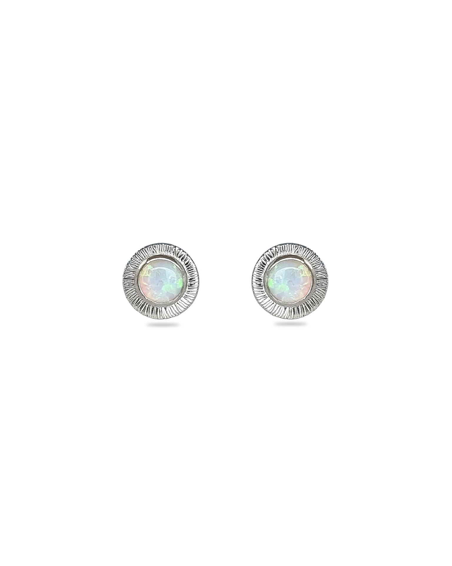A pair of handmade opal stud earrings by Emily Eliza Arlotte, featuring white opals and an engraved textured halo, on a white background.