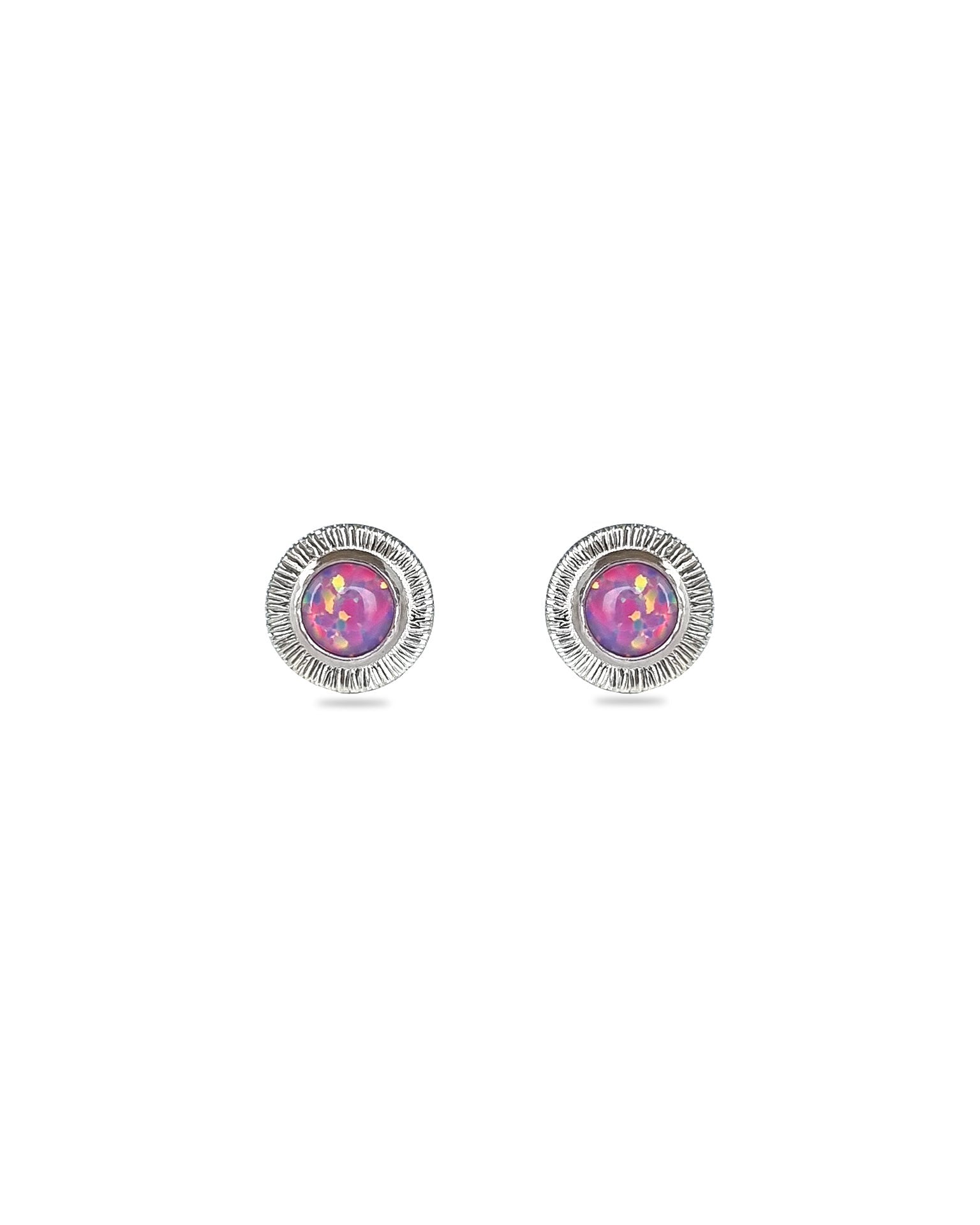 A pair of handmade opal stud earrings by Emily Eliza Arlotte, featuring violet purple opals and an engraved textured halo, on a white background.