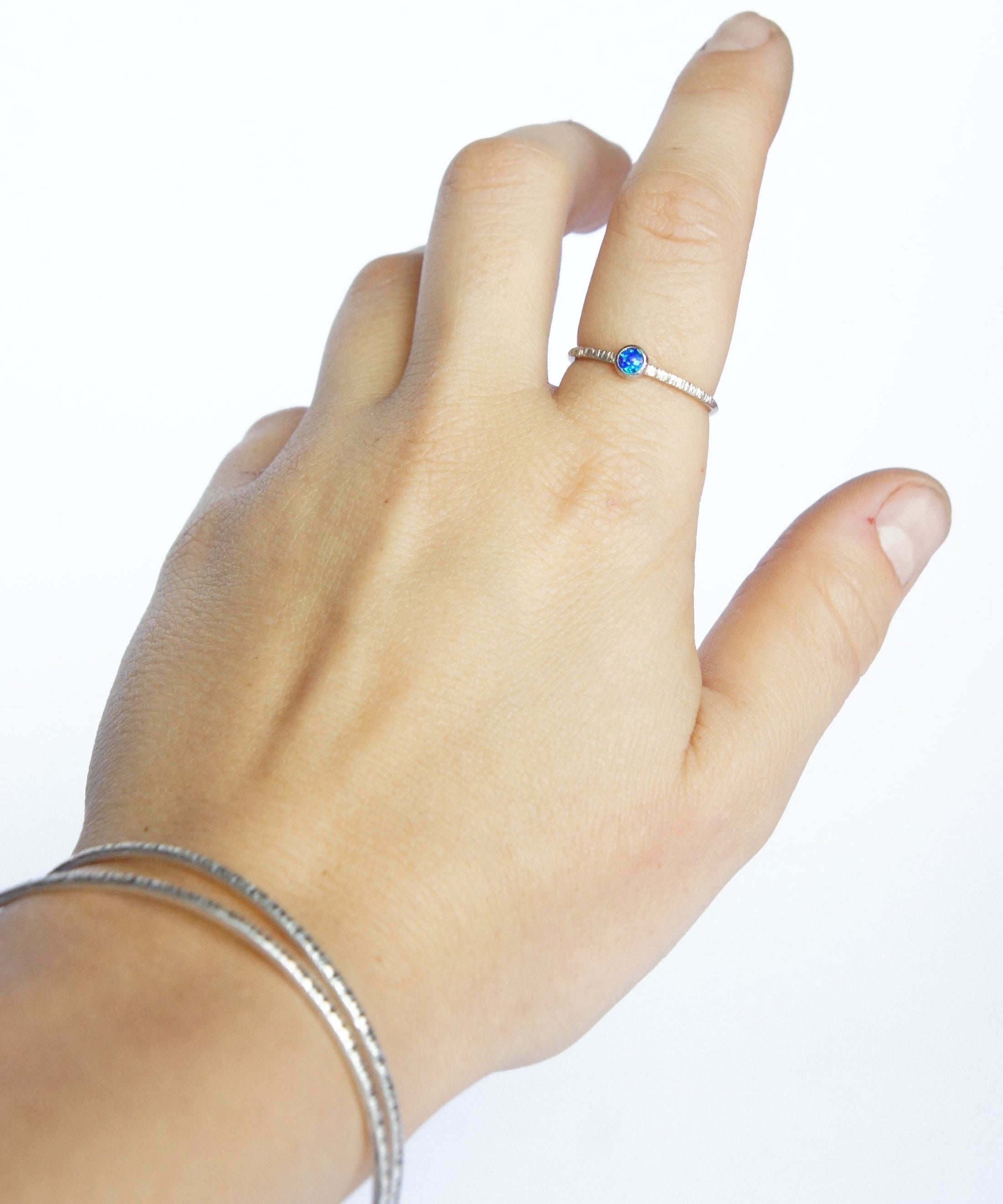 A hand on a white background is wearing a Sterling Silver and Opal Ring along with two textured bangles, handmade by Emily Eliza Arlotte in Hobart, Tasmania.