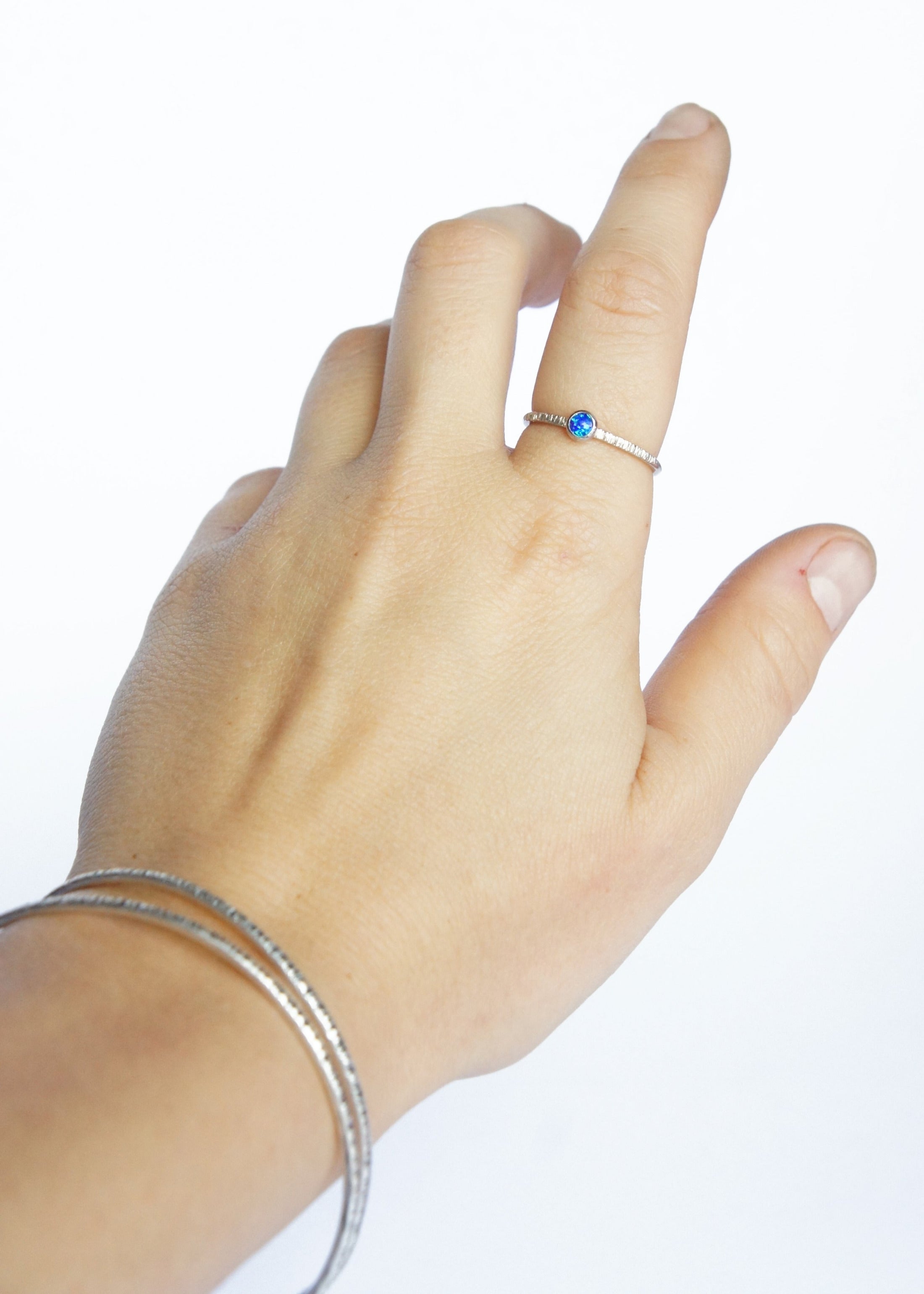A hand on a white background is wearing a Sterling Silver and Opal Ring along with two textured bangles, handmade by Emily Eliza Arlotte in Hobart, Tasmania.