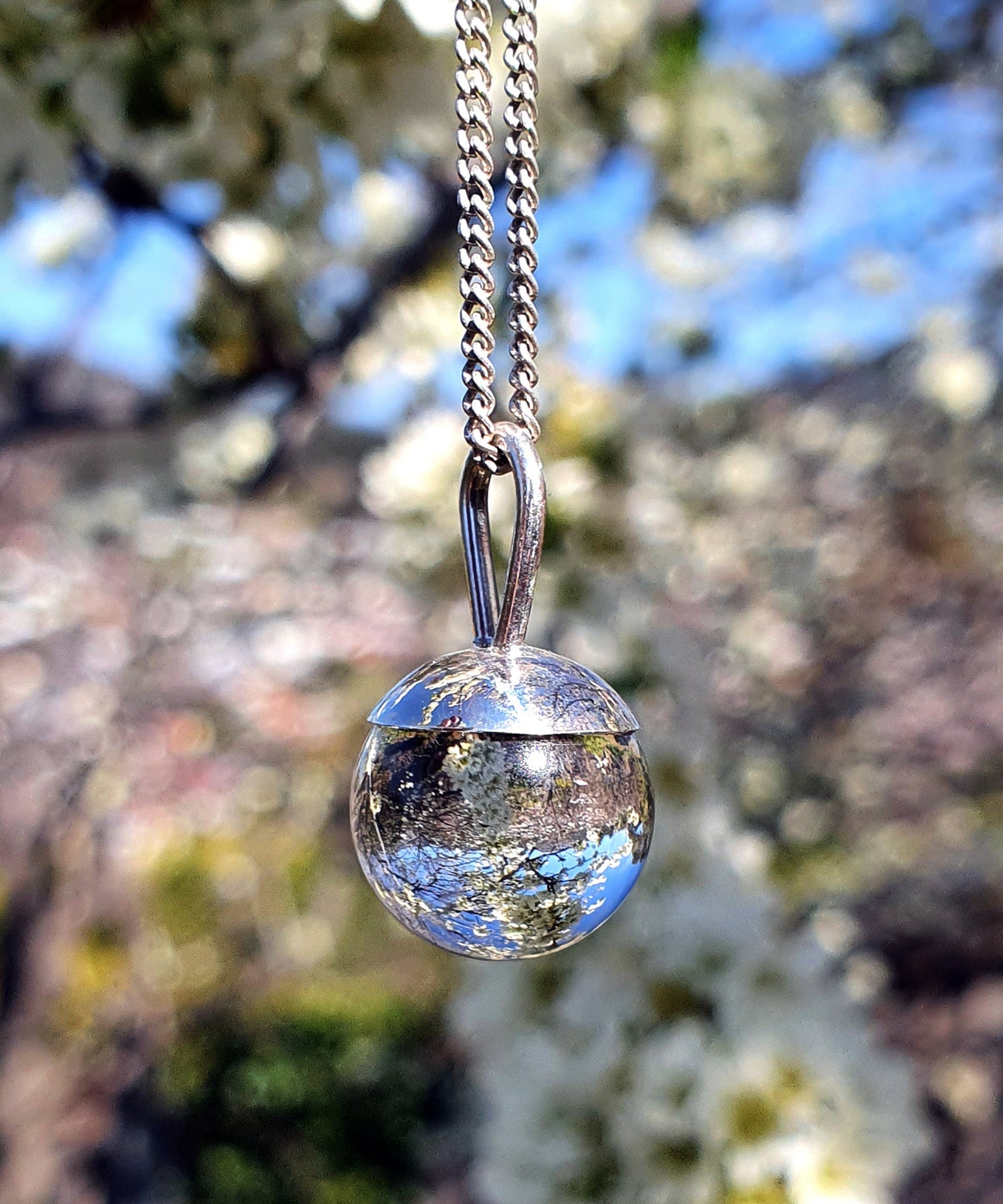 The Divination Orb Necklace hangs in front of a apple blossom tree, reflecting and inverting the image of the sky and blossoms within the orb. The sterling silver pendant necklace features a quartz crystal ball, and is handmade by Emily Eliza Arlotte in Hobart, Tasmania.