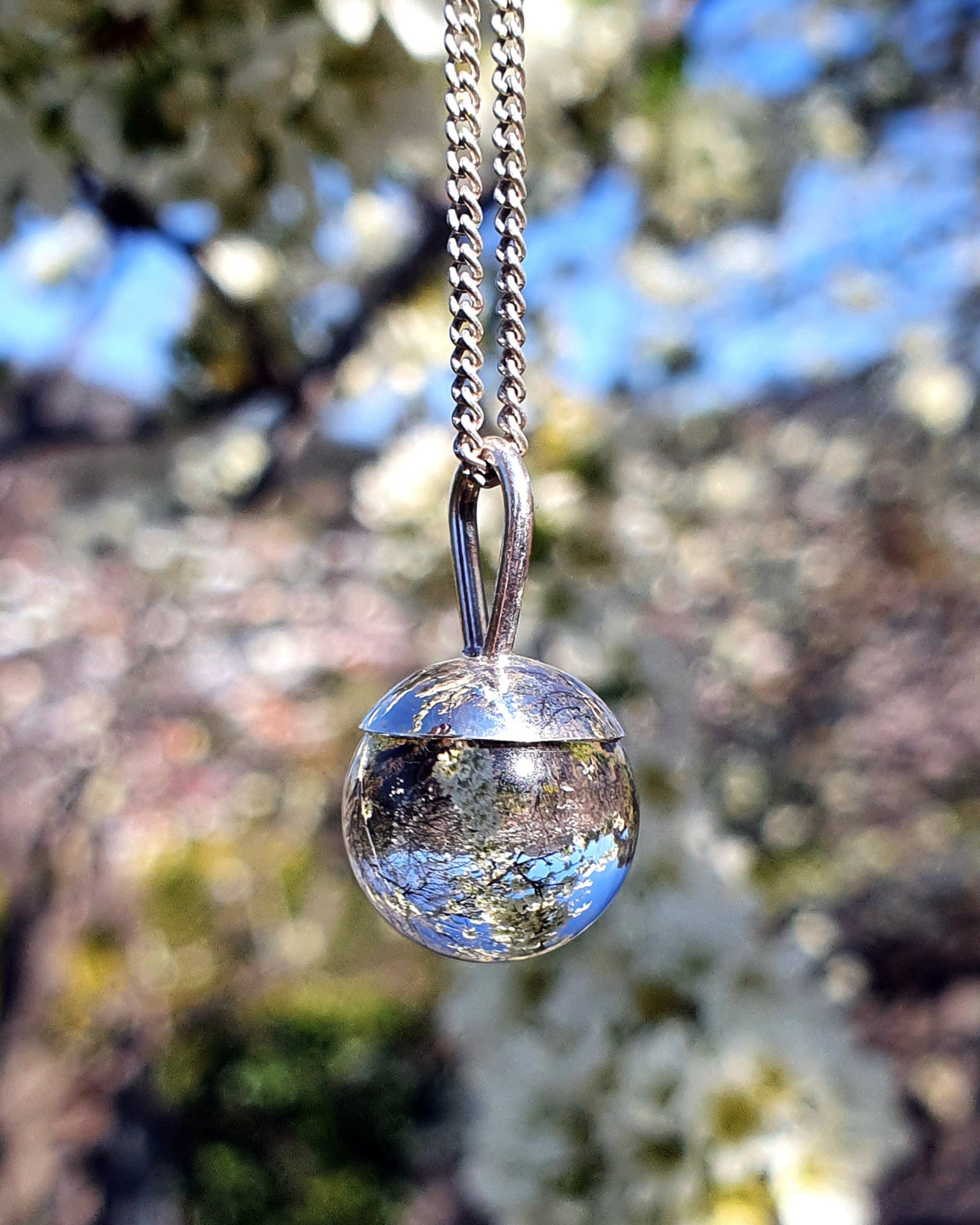 The Divination Orb Necklace hangs in front of a apple blossom tree, reflecting and inverting the image of the sky and blossoms within the orb. The sterling silver pendant necklace features a quartz crystal ball, and is handmade by Emily Eliza Arlotte in Hobart, Tasmania.