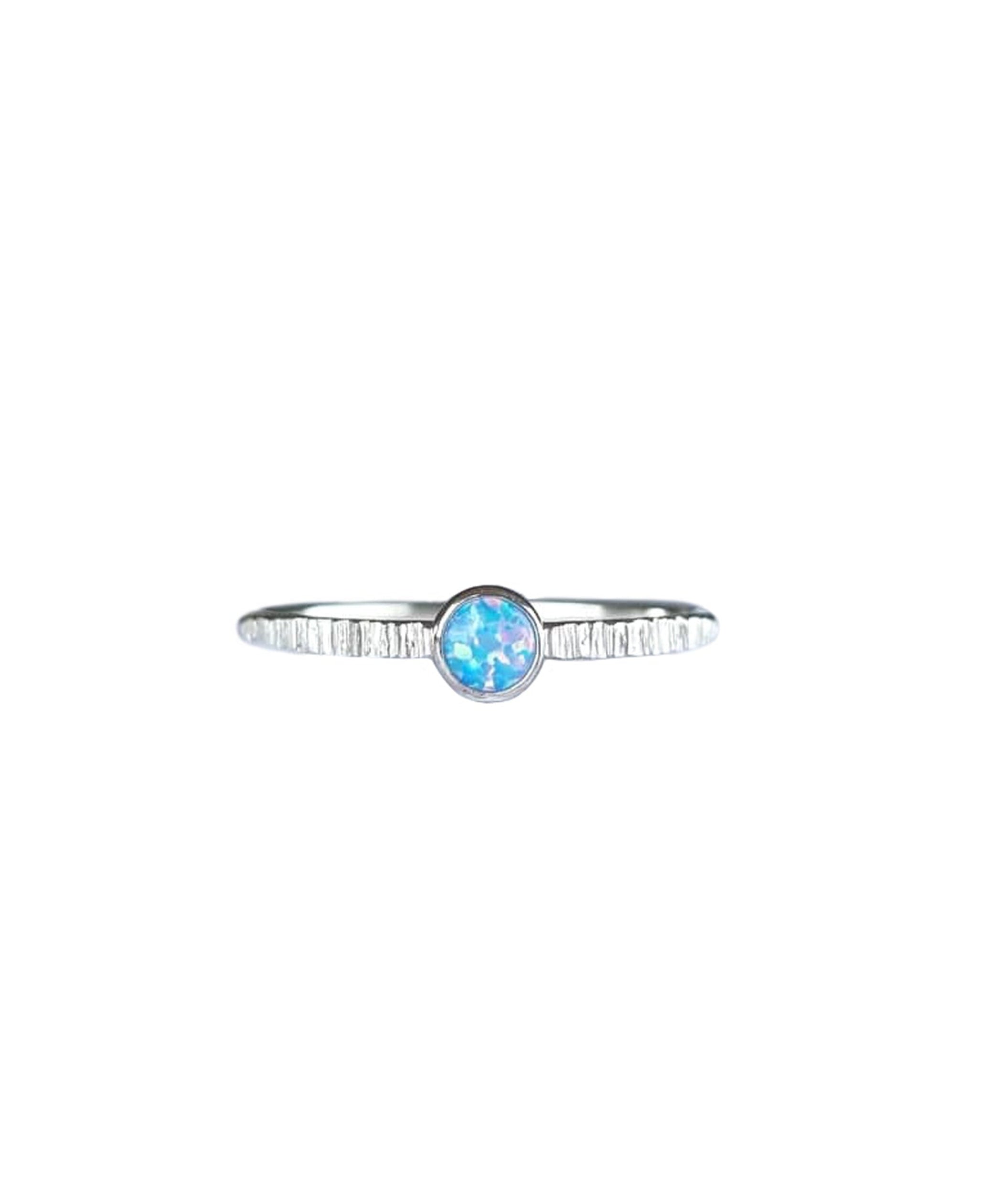 A Sterling Silver Opal Ring, with a subtle texture on a fine band sits on a background of white. The opal is a soft blue colour with shifting pinks.
