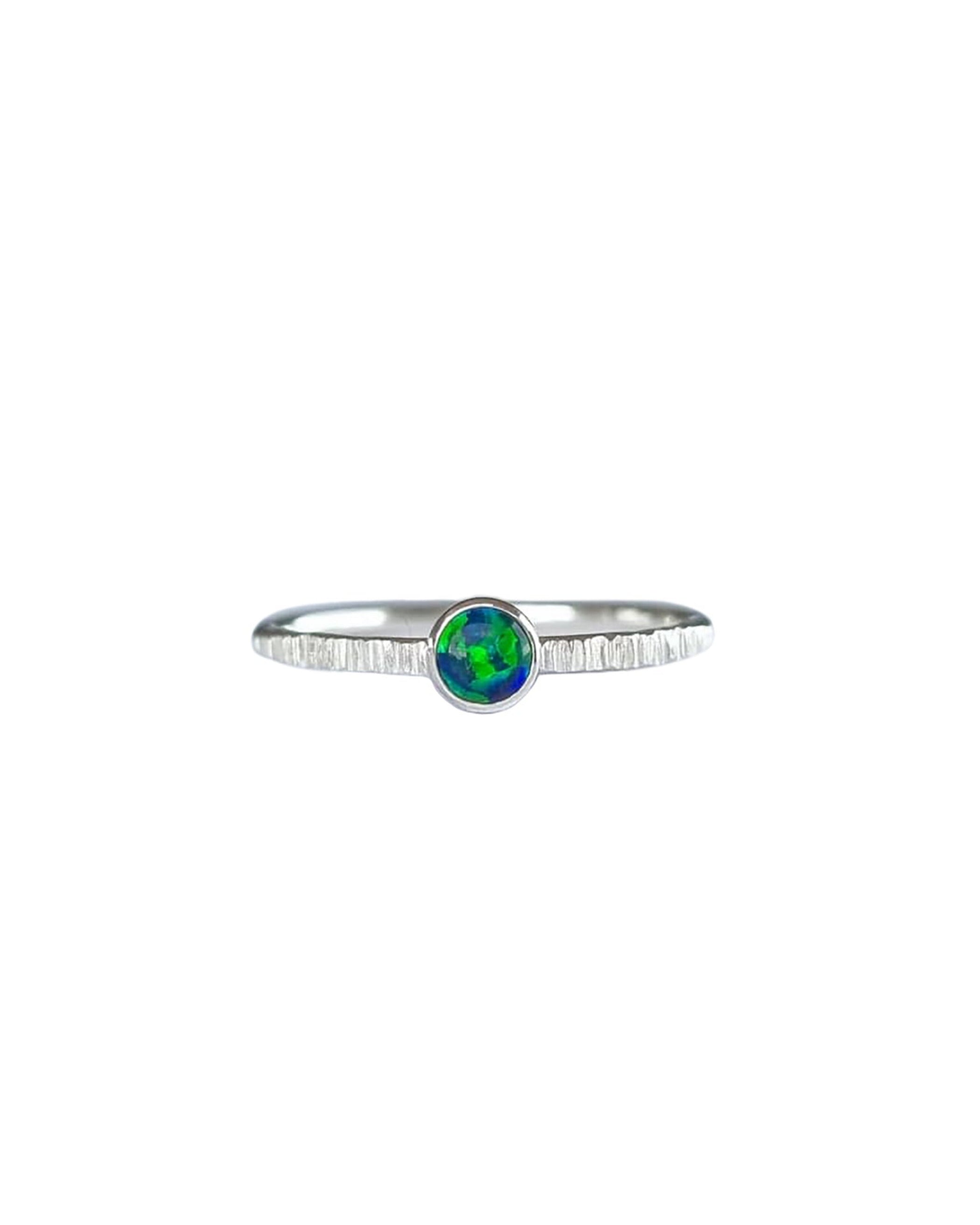 A Sterling Silver Opal Ring, with a subtle texture on a fine band sits on a background of white. The opal is a deep forest green colour with shifting dark blue flashes.