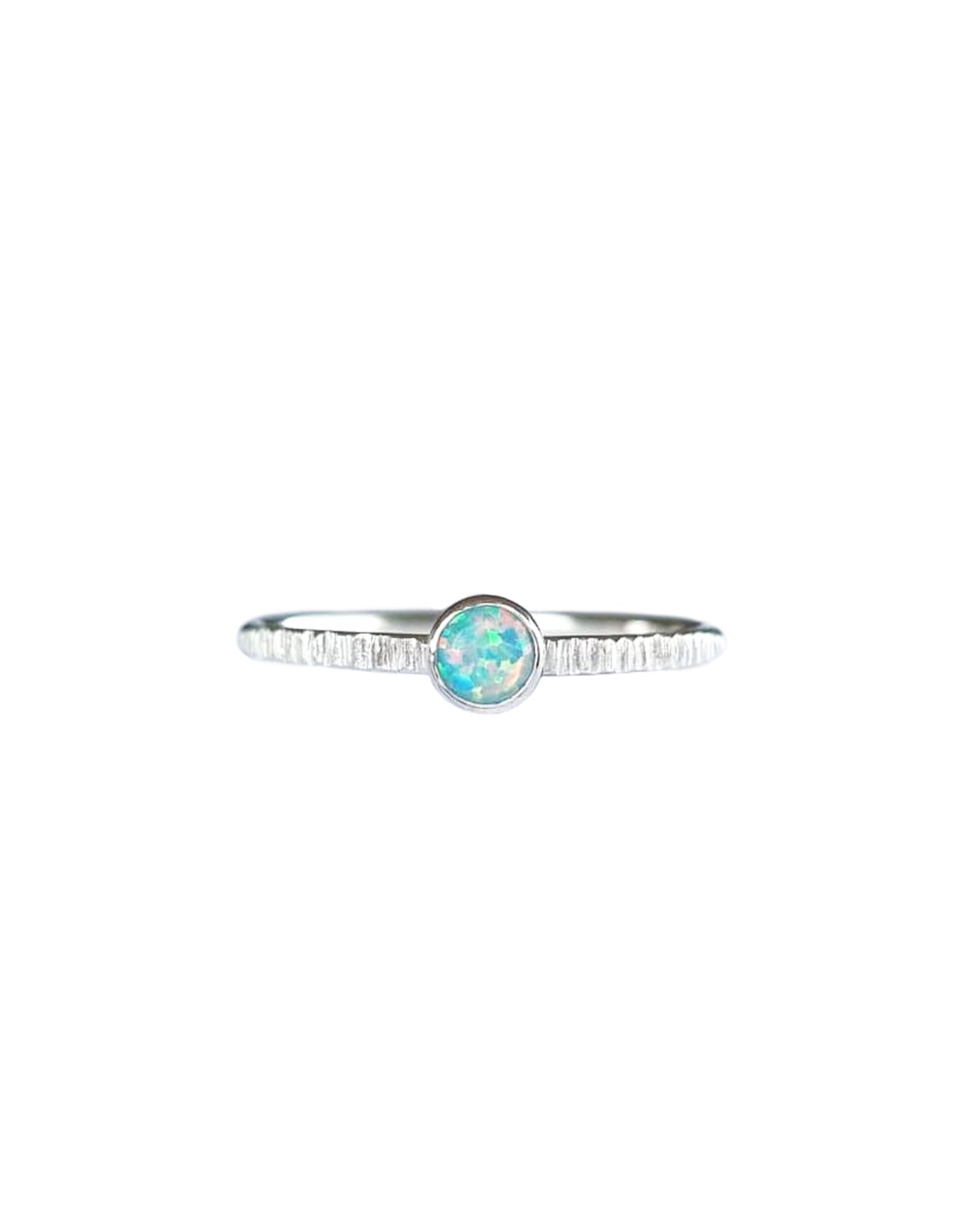 A Sterling Silver Opal Ring, with a subtle texture on a fine band sits on a background of white. The opal is a soft green colour of shifting pinks and blues.