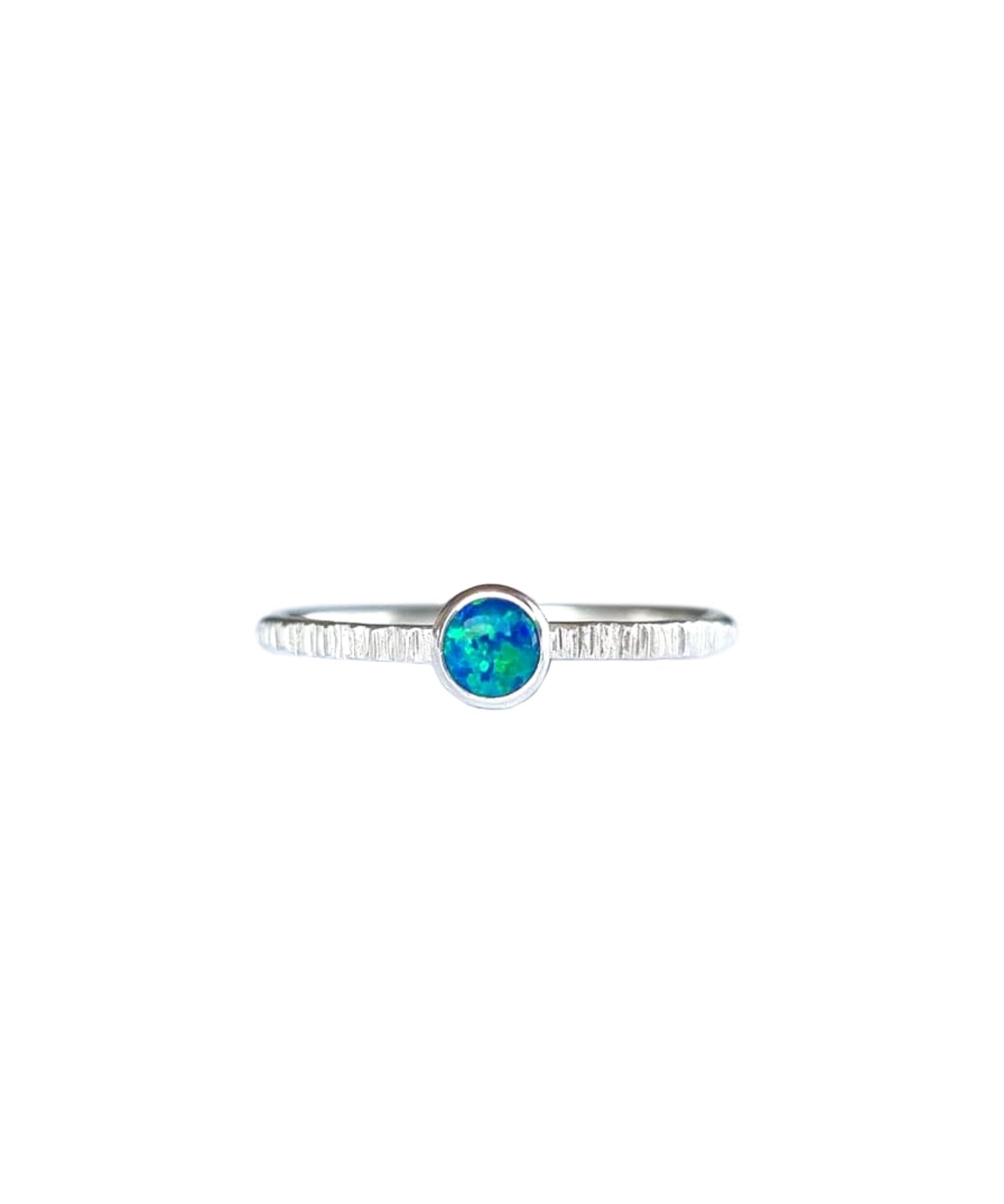 A Sterling Silver Opal Ring, with a subtle texture on a fine band sits on a background of white. The opal is an ocean green colour with shifting blue flashes.
