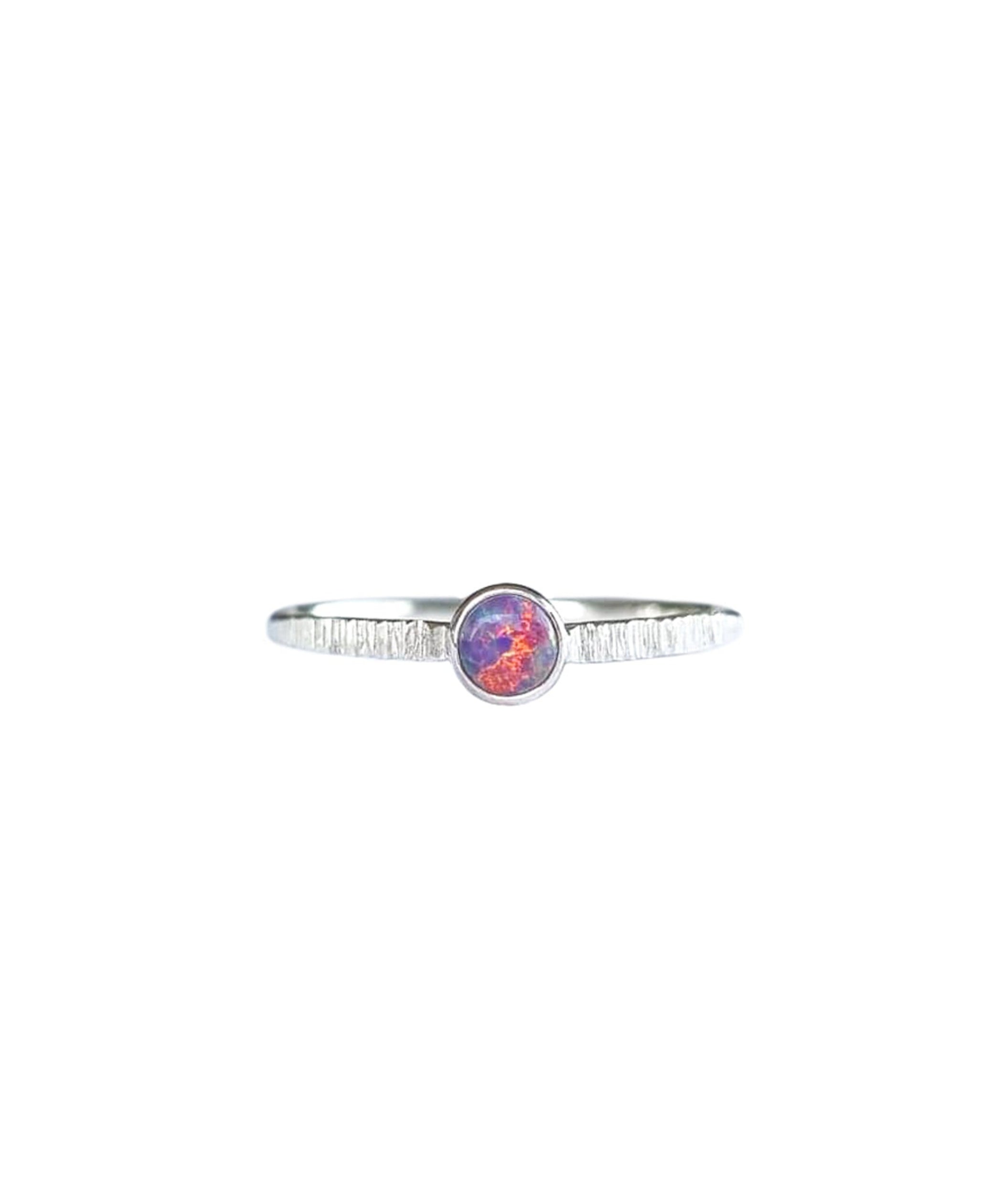 A Sterling Silver Opal Ring, with a subtle texture on a fine band sits on a background of white. The opal is a violet purple colour of shifting pinks and oranges.
