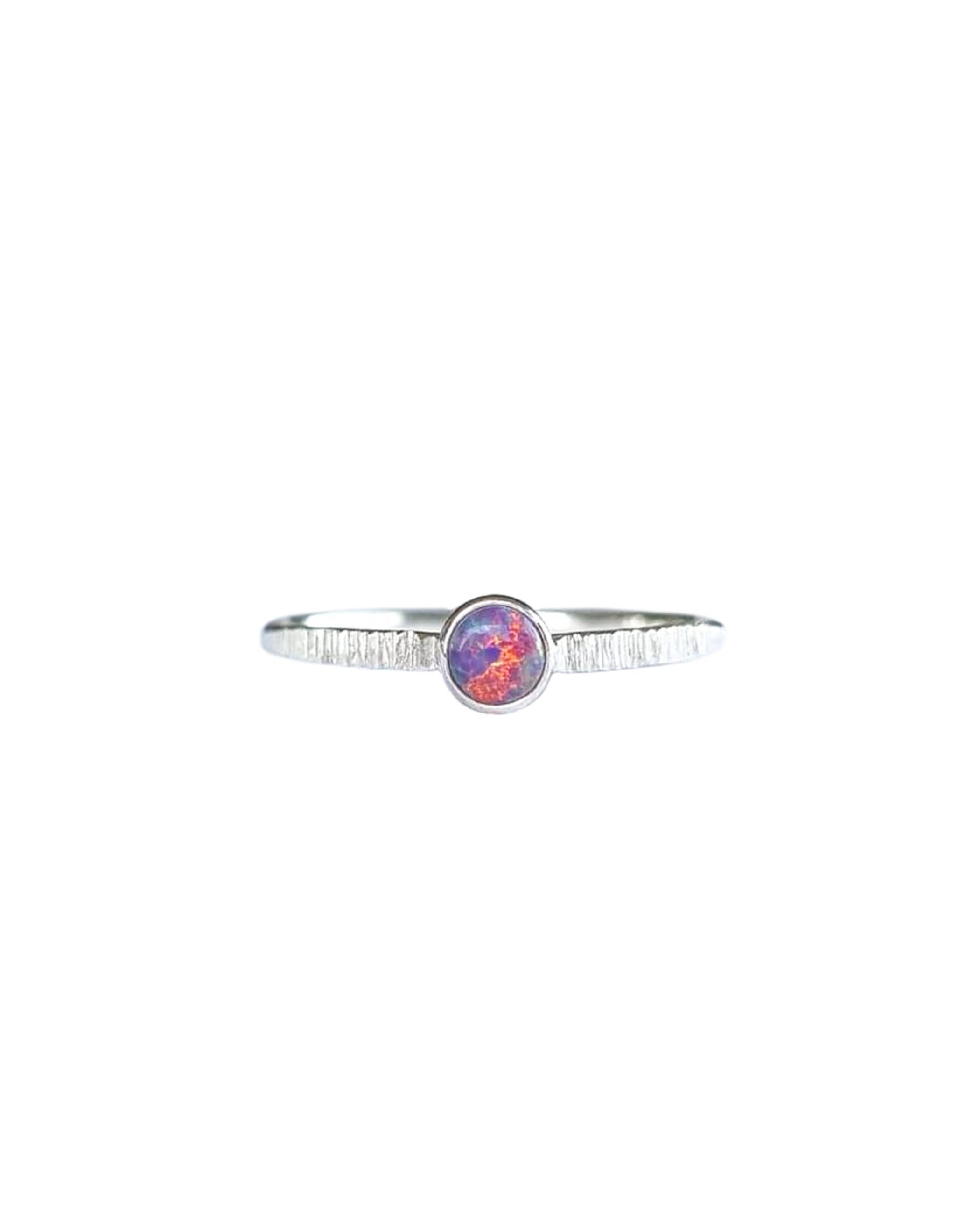 A Sterling Silver Opal Ring, with a subtle texture on a fine band sits on a background of white. The opal is a violet purple colour of shifting pinks and oranges.