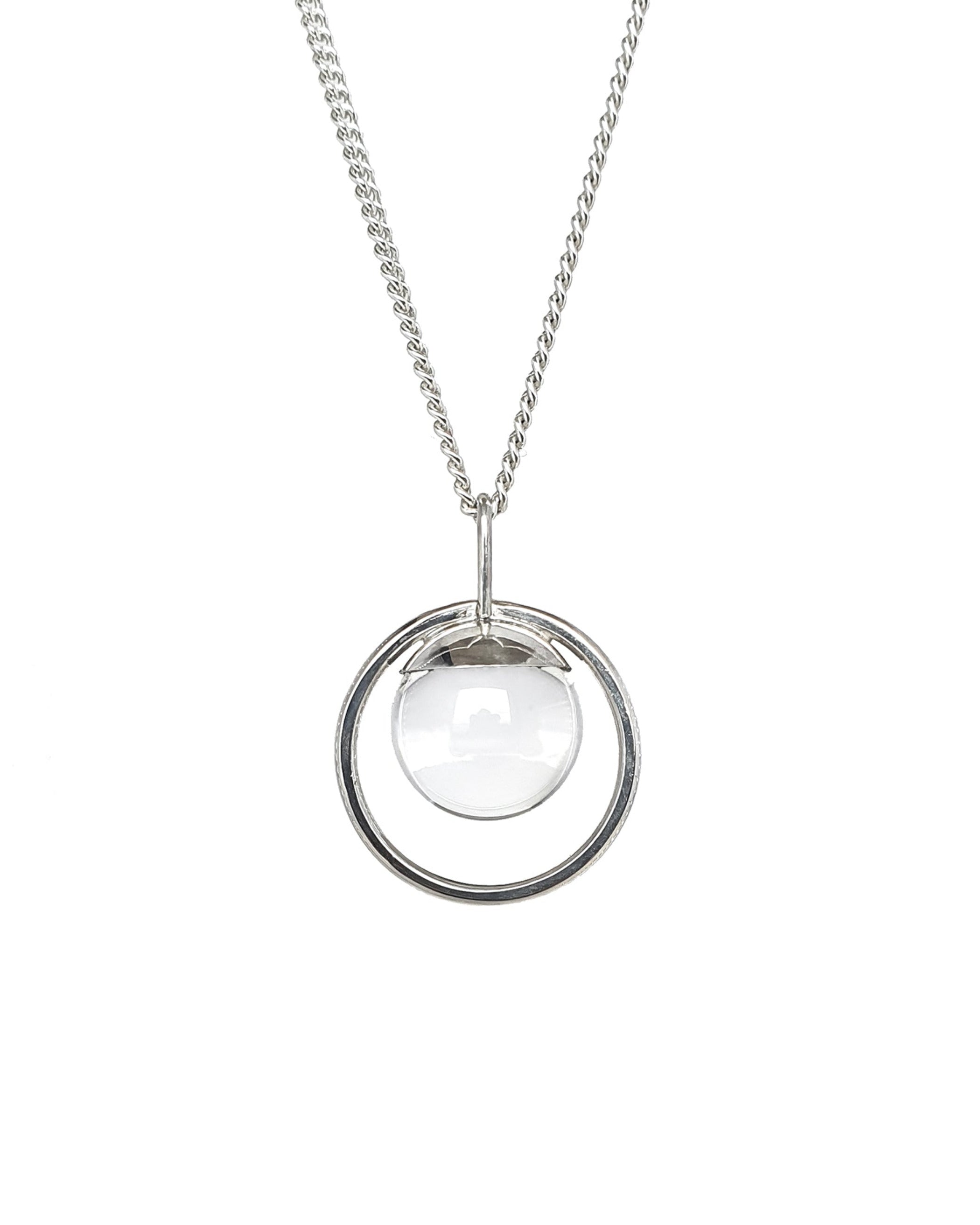 The Divination Sterling Silver Necklace handmade by Emily Eliza Arlotte in Hobart, Tasmania, hangs on a white background. It features a quartz sphere, or crystal ball, hung inside a circle of silver.
