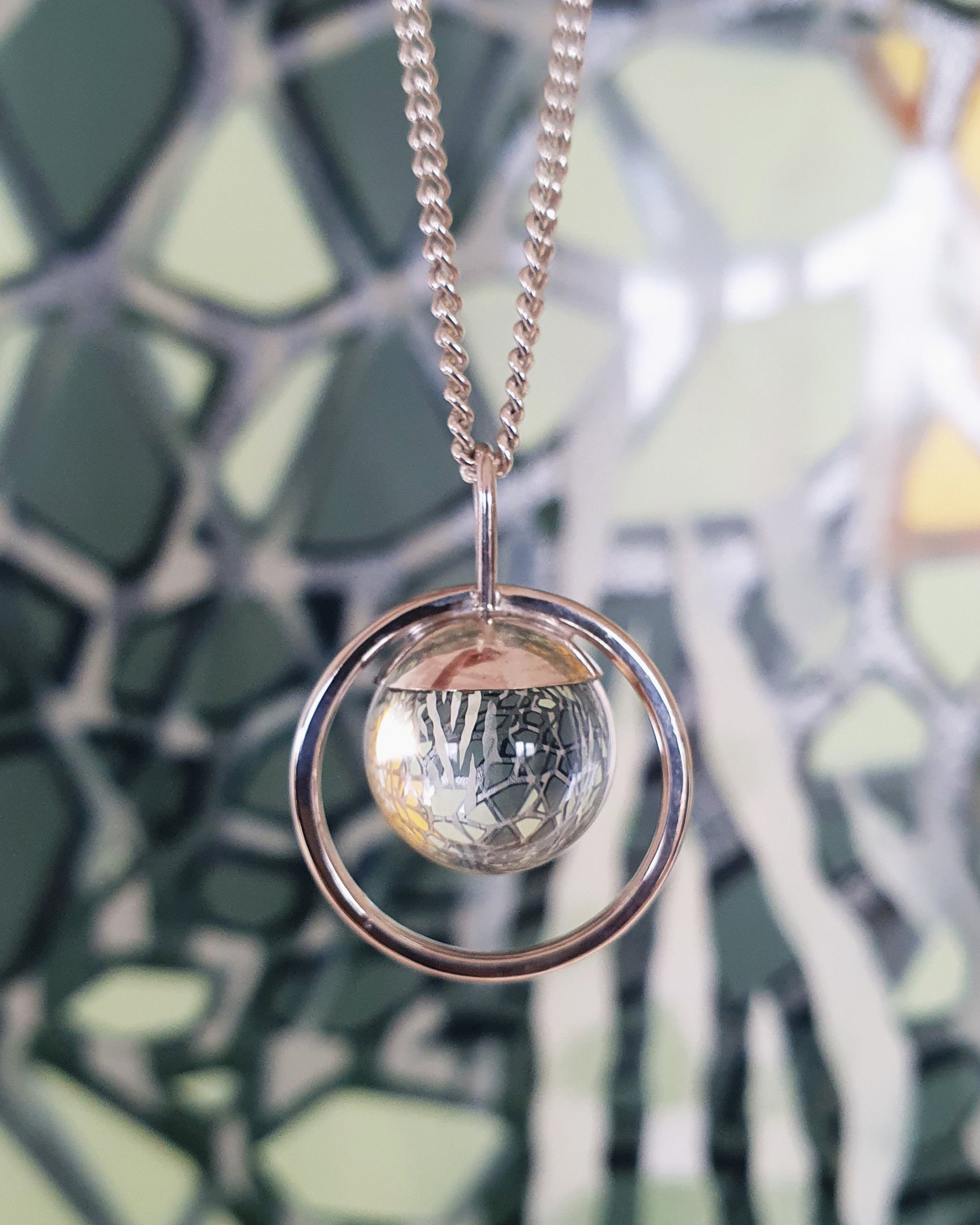 The Divination Necklace, handmade by Emily Eliza Arlotte, features a clear quartz sphere reflecting and inverting the image of an artwork by Jamin Kluss behind it.