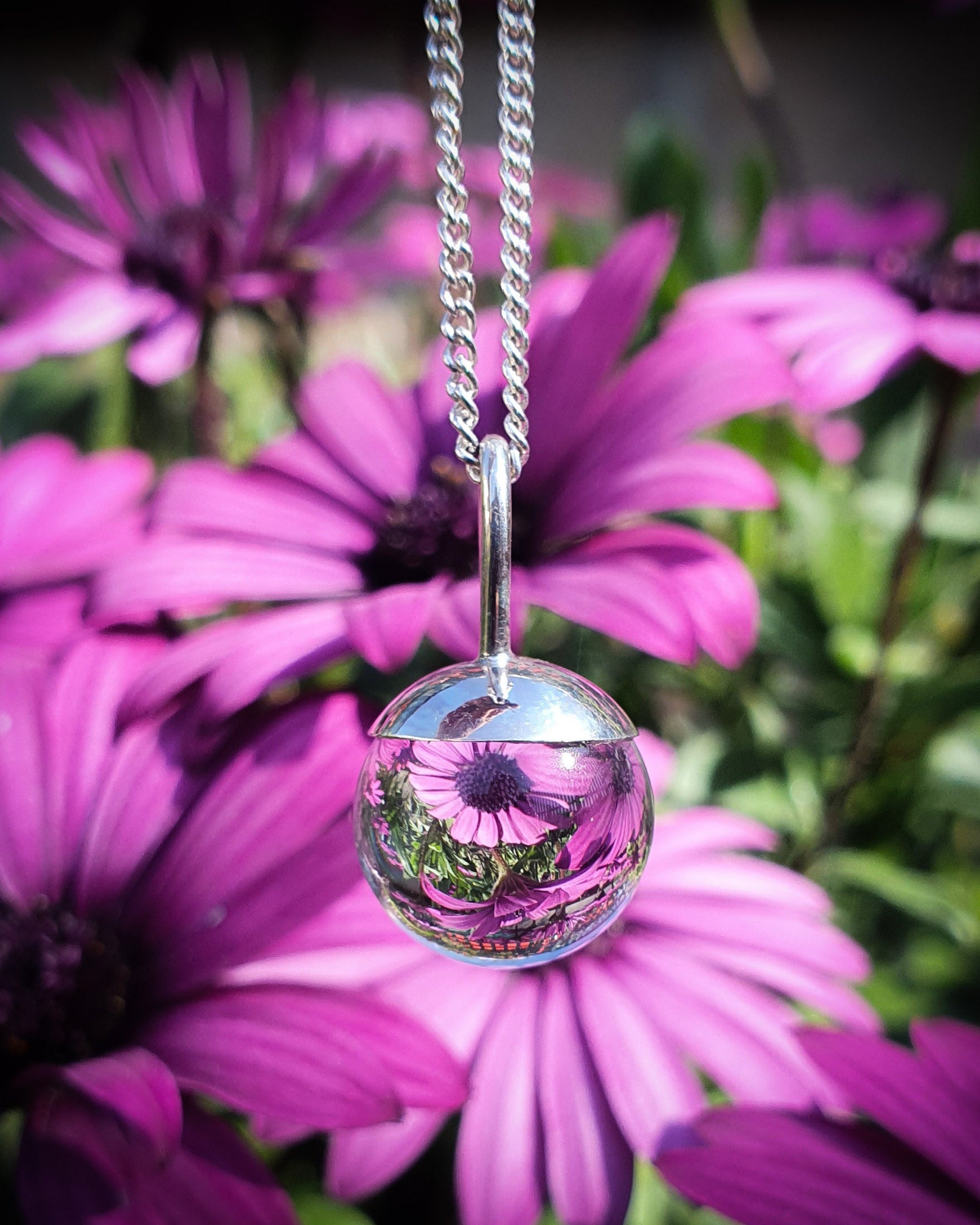 The Divination Orb Necklace hangs in front of a flower garden, reflecting and inverting the image of purple daisy flowers. The sterling silver pendant necklace features a quartz crystal ball, and is handmade by Emily Eliza Arlotte in Hobart, Tasmania.