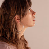The Venus Hoop Earrings, which are an oversized hoop in the shape of the feminist symbol, worn by a person wearing pink on a soft pink background. The earrings are handmade by Emily Eliza Arlotte in Hobart, Tasmania 