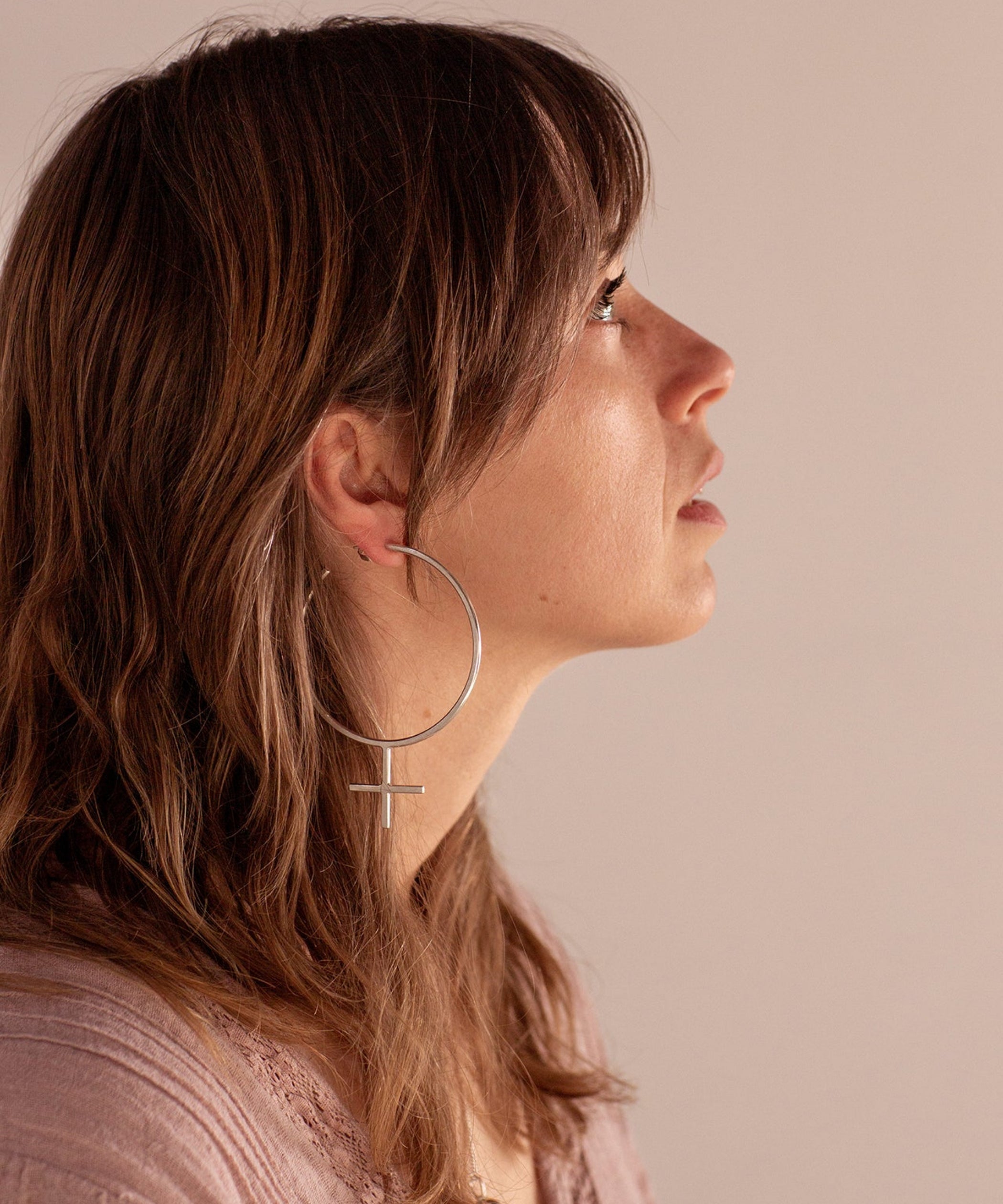 The Venus Hoop Earrings, which are an oversized hoop in the shape of the feminist symbol, worn by a person wearing pink on a soft pink background. The earrings are handmade by Emily Eliza Arlotte in Hobart, Tasmania 