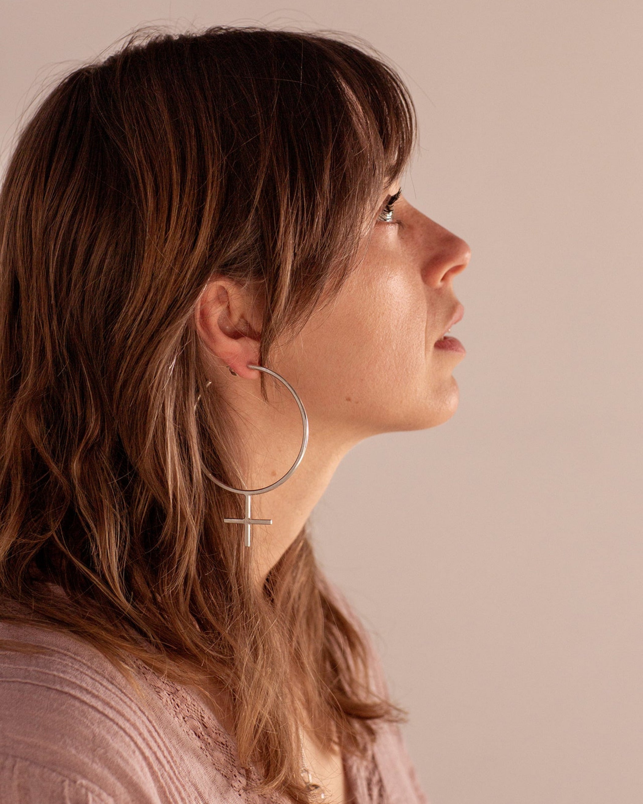 The Venus Hoop Earrings, which are an oversized hoop in the shape of the feminist symbol, worn by a person wearing pink on a soft pink background. The earrings are handmade by Emily Eliza Arlotte in Hobart, Tasmania 